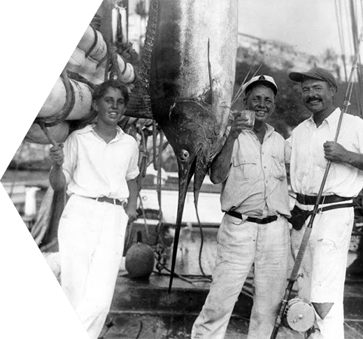 Old black and white photo of a young Ernest Hemingway standing next to a marlin that has been hung from its tail in Key West, FL