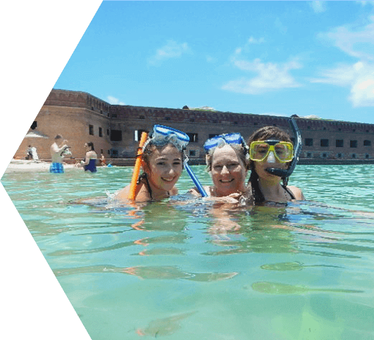 A mother and her two daughters in the ocean smiling with their snorkeling masks propped on top of their heads with Ft. Jefferson and the Dry Tortugas National Park in the background