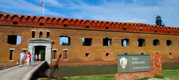 guests on a walkway above a moat that connects to the entrance to Fort Jefferson, a large brick structure, and to the right is a sign that reads ‘Fort Jefferson Dry Tortugas National Park’ mounted on brick