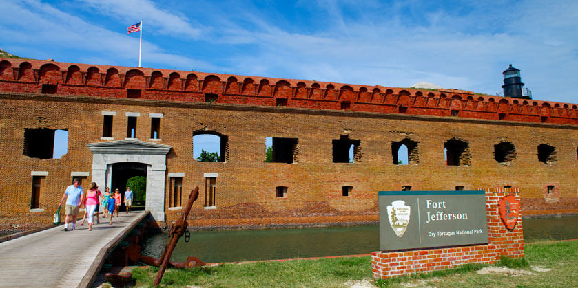 guests on a walkway above a moat that connects to the entrance to Fort Jefferson, a large brick structure, and to the right is a sign that reads ‘Fort Jefferson Dry Tortugas National Park’ mounted on brick